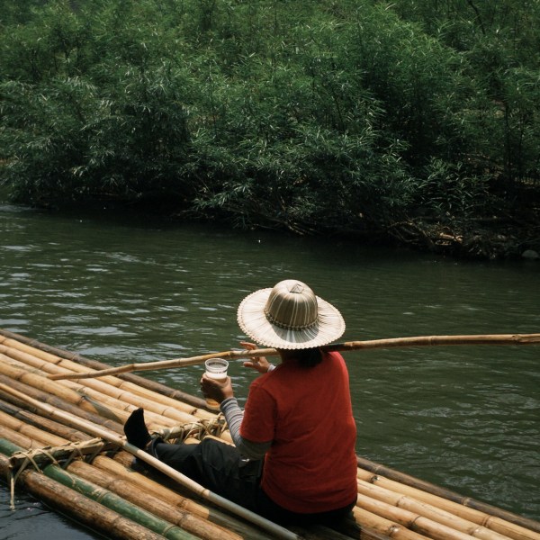 Person in straw hat sits on bamboo raft on a river, holding a cup.
