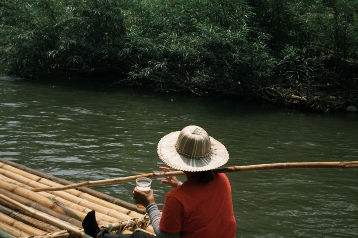 Person in straw hat sits on bamboo raft on a river, holding a cup.