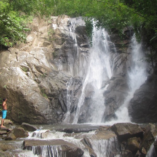 Person standing near a waterfall surrounded by rocks and greenery.