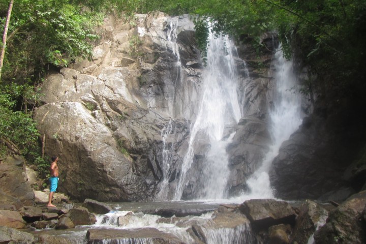 Person standing near a waterfall surrounded by rocks and greenery.
