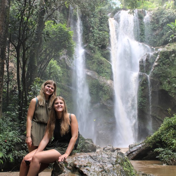 Two people sit on rocks near a waterfall in a lush forest.